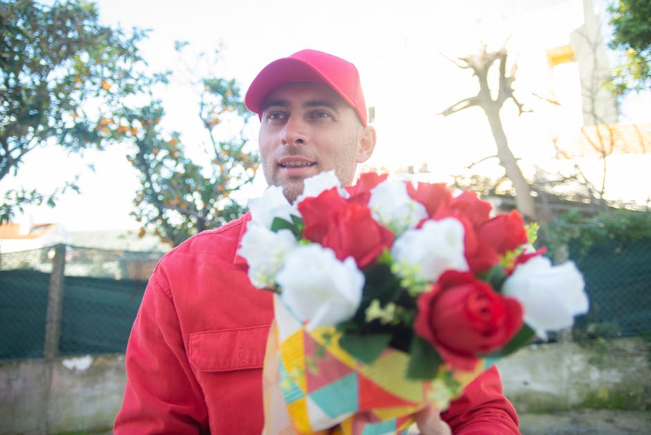 A male flower delivery courier dressed in a red uniform and matching cap is holding a vibrant bouquet of fresh flowers, featuring red and white roses with green foliage, wrapped in colorful paper. He