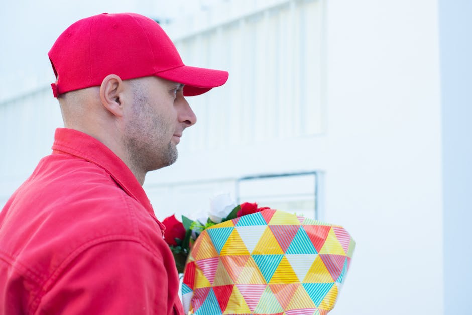 A delivery person dressed in a red uniform and matching cap is holding a large, colorful floral gift wrapped in vibrant geometric patterned paper with shades of yellow, blue, red, and orange. The bouq