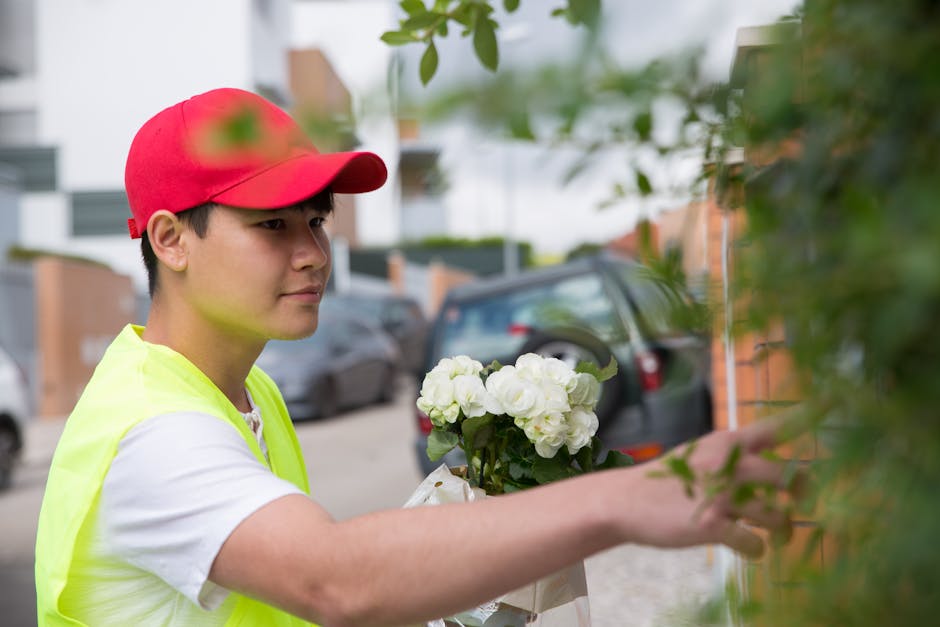 A young male florist wearing a red cap and a neon yellow vest delivers a floral gift outside a residential building, holding a bouquet of white hydrangeas wrapped in clear plastic. The arrangement app