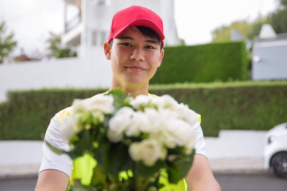 A young man wearing a red cap and a high-visibility safety vest is standing outdoors, holding a bouquet of white flowers with green leaves. The bouquet appears fresh with slightly textured petals and